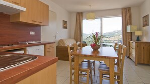 A modern kitchen with wooden cabinets, a dining table with chairs, and a view of the outdoors through large windows. - Madame Vacances Residence La Marquisié (Trebas)