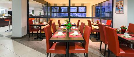 A modern dining area with red chairs and tables, large windows, and a view of the cityscape.