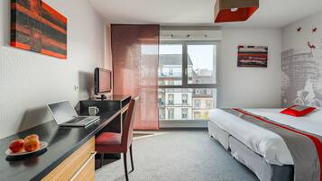 A modern hotel room with a desk, laptop, and two apples on a plate, a bed with a red and grey bedspread, and a cityscape view through the window.