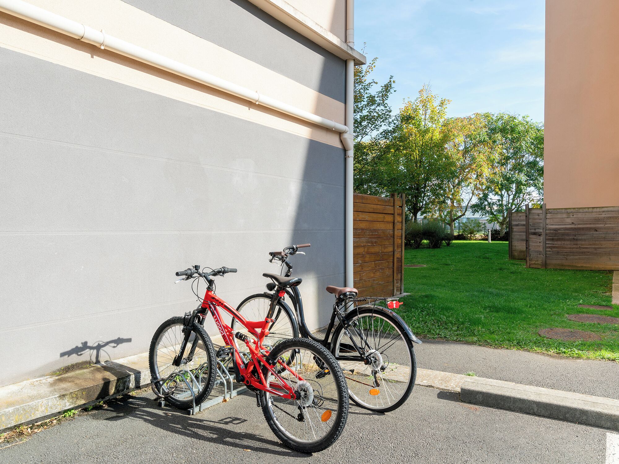 Two bicycles parked against a building wall in a well-maintained urban area with a grassy lawn and trees in the background.