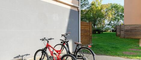 Dos bicicletas estacionadas junto a una pared de edificio en una zona urbana bien cuidada, con césped y árboles al fondo.