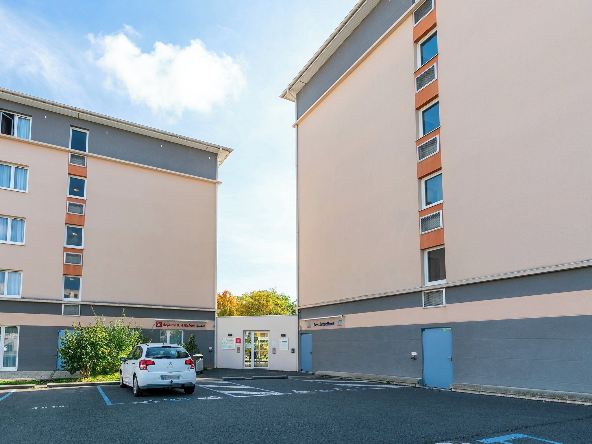 A parking lot with a white car, two buildings with beige and grey facades, and a sign for 'Les Estudines'.