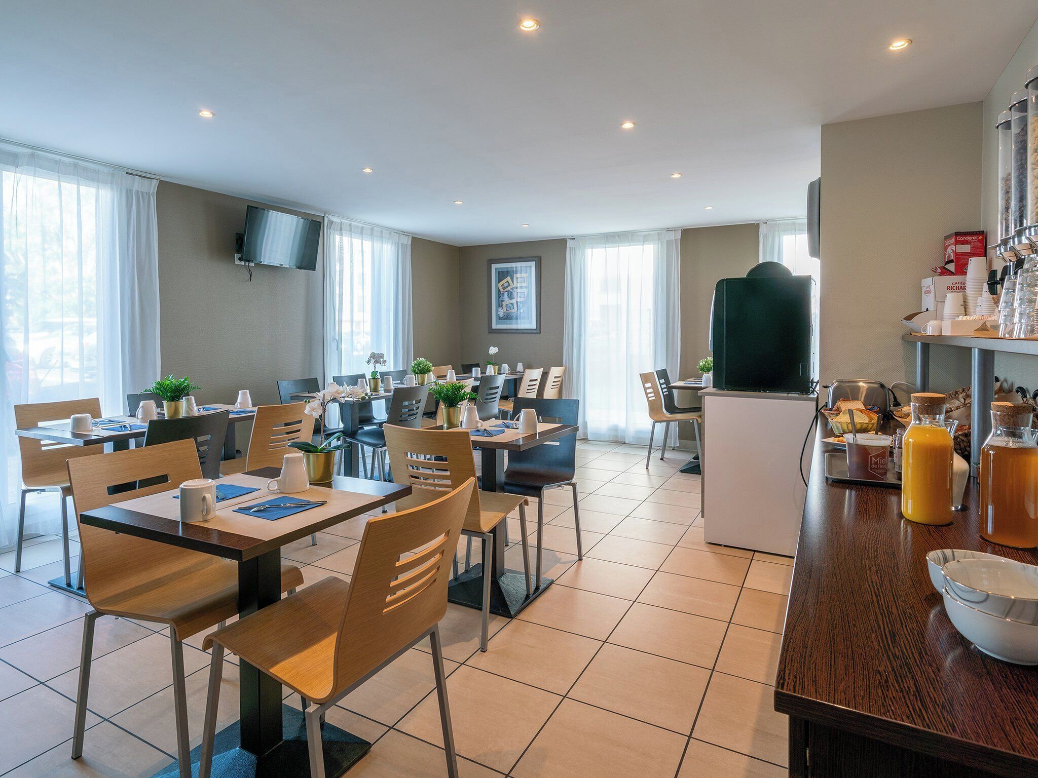A modern hotel dining area with multiple tables and chairs, a coffee station, and a television mounted on the wall.