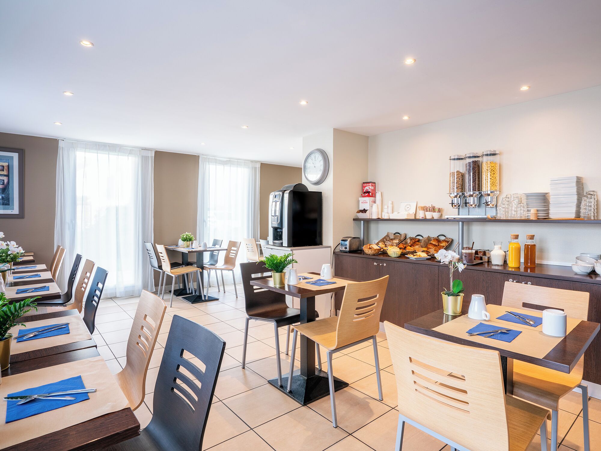 A modern dining area with multiple tables and chairs, a kitchen area with a coffee machine, and a clock on the wall.