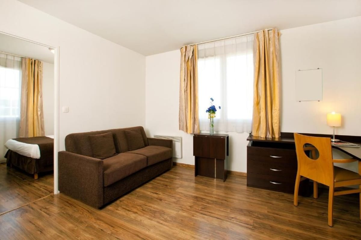 A room with a brown sofa, a wooden chair, a small desk, a lamp, and a window with curtains.