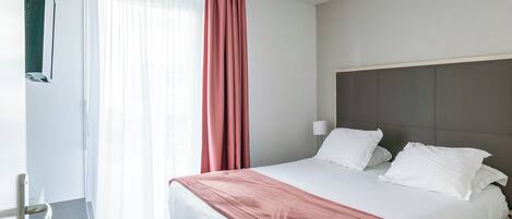 Una habitación de hotel moderna con una cama grande, una mesita de noche, una lámpara y una ventana con cortinas.