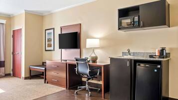 A hotel room with a wooden desk, a black office chair, a flat-screen TV, a microwave, a coffee maker, and a refrigerator.