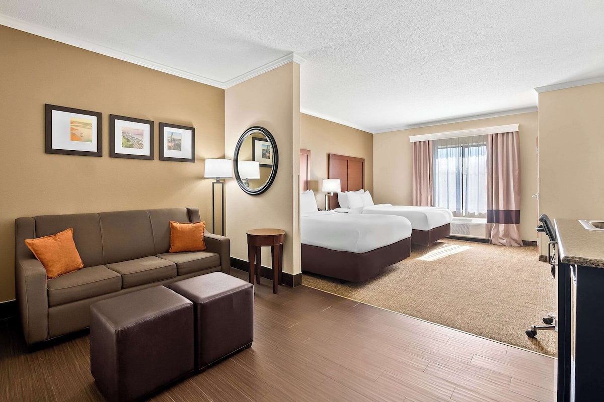 A hotel room with a sofa, two beds, a small table, and framed pictures on the wall.