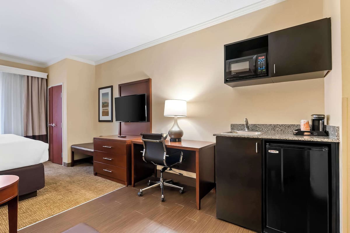 A hotel room with a bed, desk, chair, TV, lamp, microwave, and refrigerator.