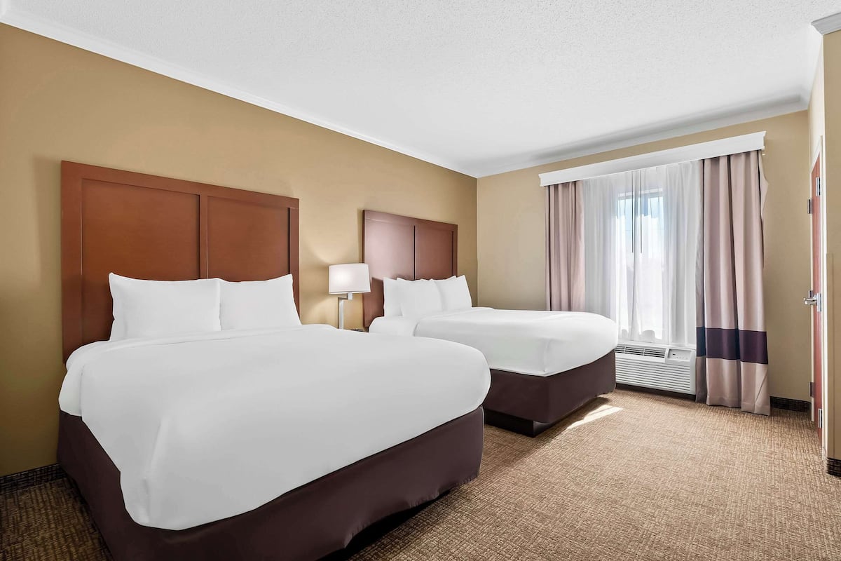 A hotel room with two beds, beige walls, a large window with curtains, and a carpeted floor.