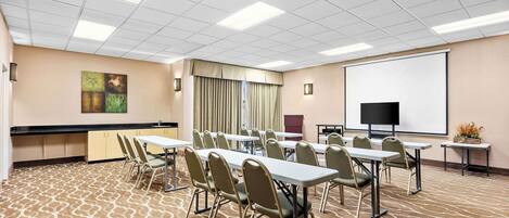 Un salón de conferencias con varias mesas y sillas, un proyector, una pantalla y un ventanal con cortinas.