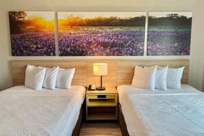 A hotel room with two beds, a nightstand with a lamp, and a wall-mounted painting of a field. - Days Inn & Suites by Wyndham Houston / West Energy Corridor (Houston)