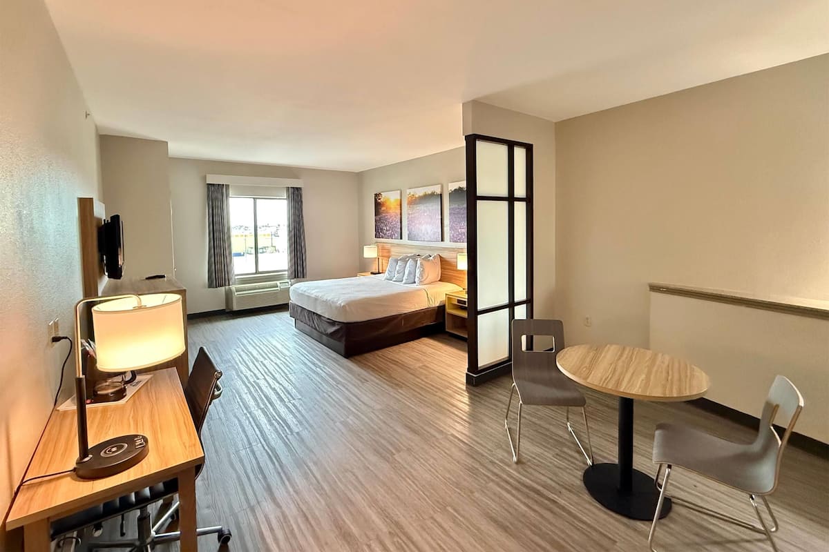 Una habitación de hotel con cama, mesita de noche con lámpara, escritorio con silla, una mesa redonda con sillas y un ventanal con cortinas.