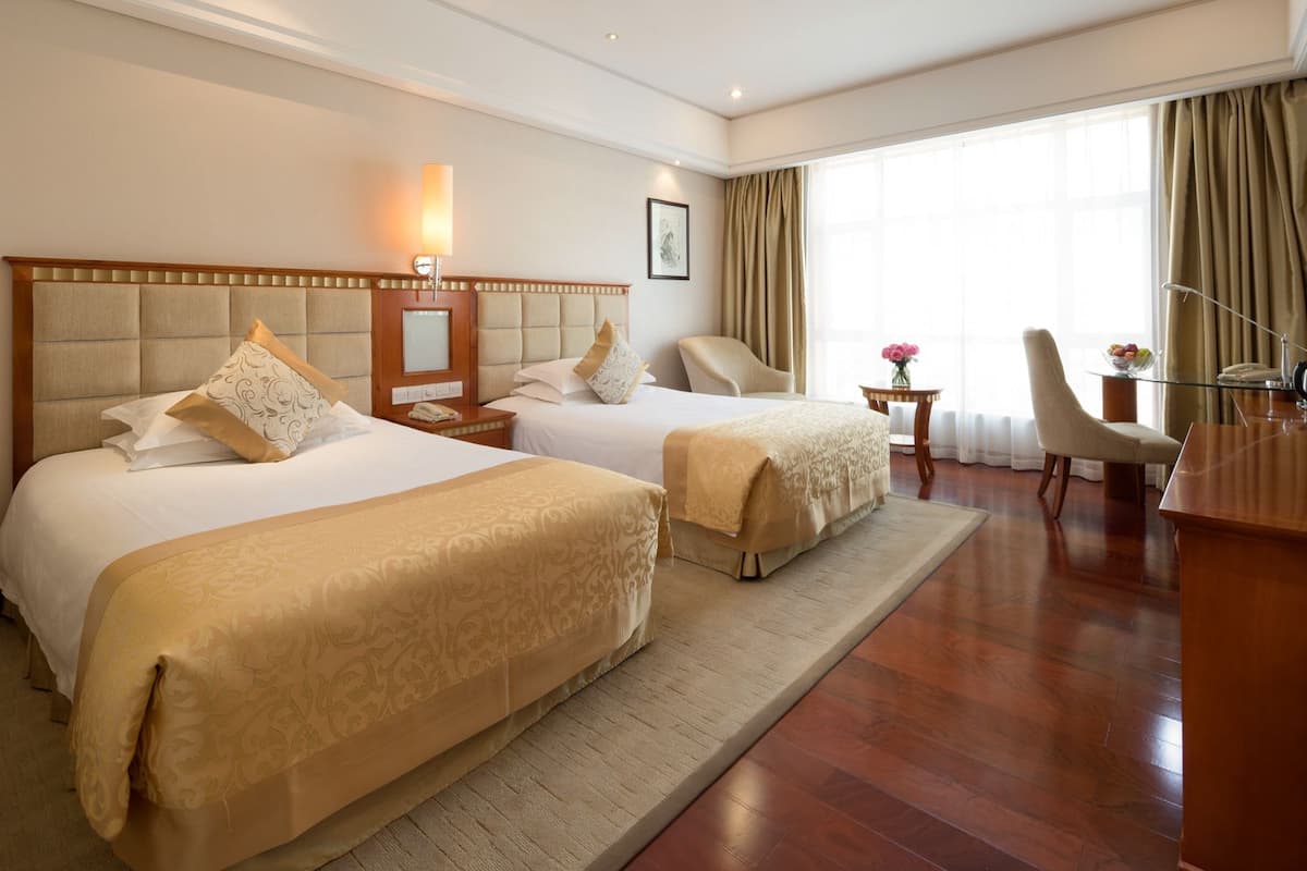 A hotel room with two beds, a desk, a chair, a small table with a vase of flowers, and a large window with curtains.