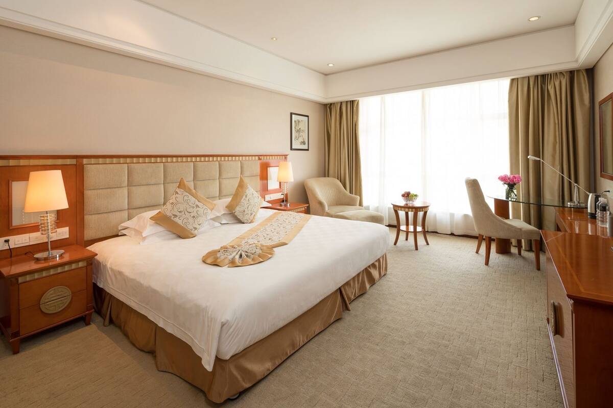 A hotel room with a large bed, a desk, two chairs, a small table, and a window with curtains.