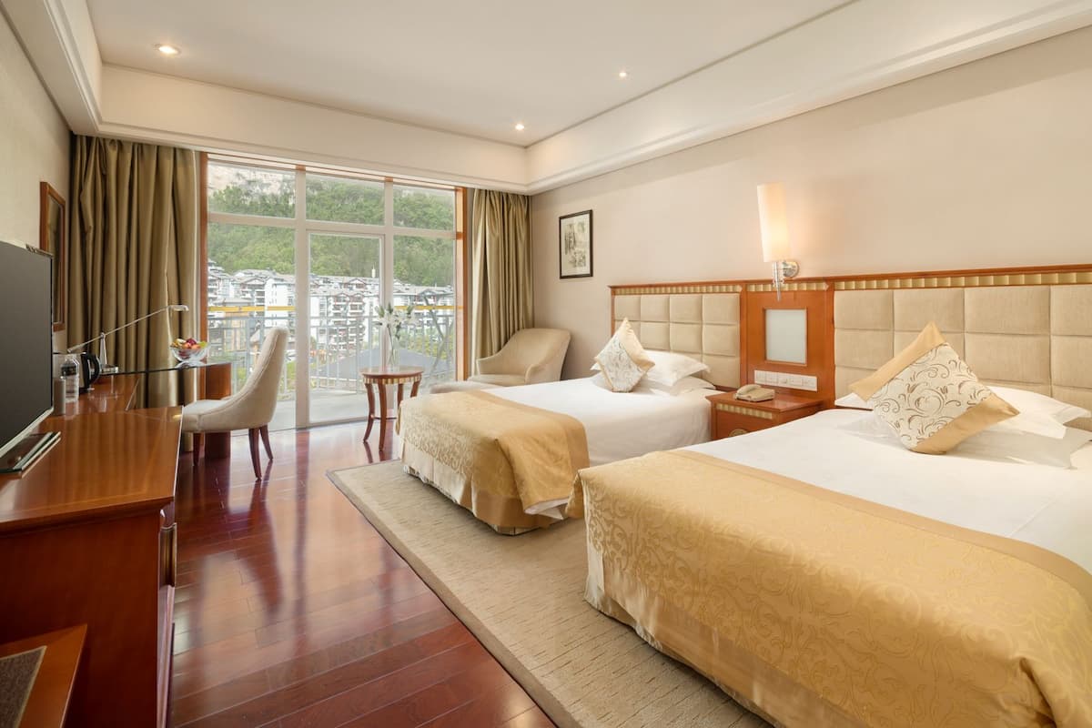 A hotel room with two beds, a desk, a chair, and a large window offering a view of buildings and trees.