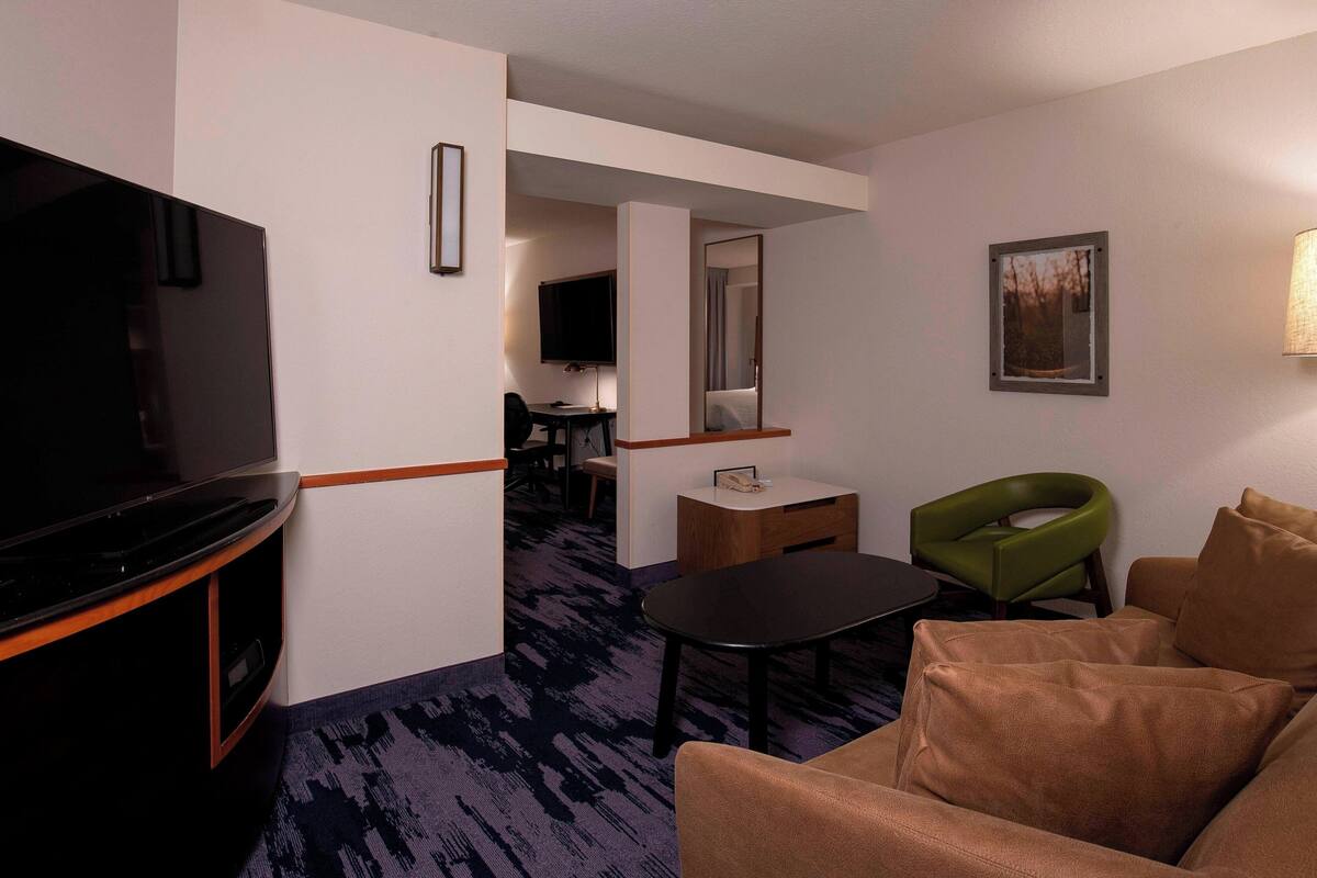 A hotel room with a flat-screen TV, a sofa, a green chair, a small table, and a mirror.