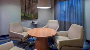 A hotel room with a round wooden table, two beige armchairs, a large painting of a forest, a ceiling light, and a window with curtains. - Fairfield Inn & Suites by Marriott Venice (Venice)
