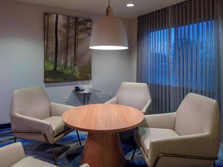 Quarto de hotel com uma mesa redonda de madeira, duas poltronas bege, uma grande pintura de uma floresta, uma luminária no teto e uma janela com cortinas.