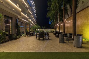 Un patio al aire libre con asientos y mesas, rodeado de palmeras y un edificio con ventanas iluminadas.