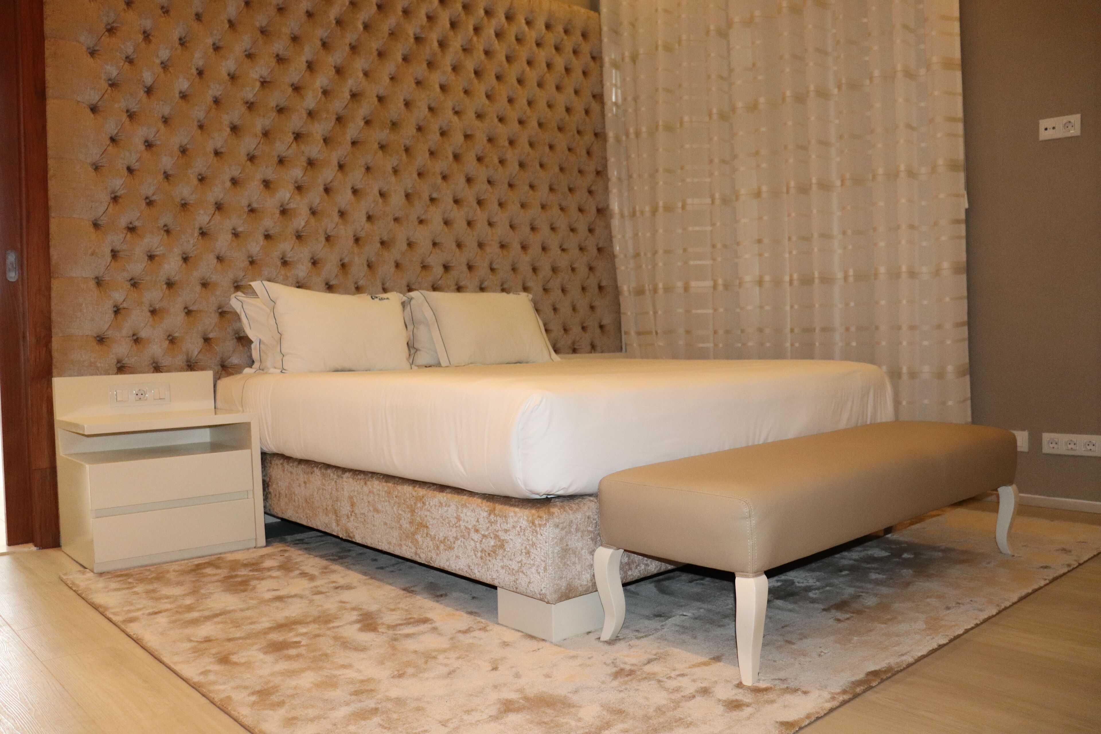 Une chambre à coucher avec une tête de lit capitonnée, un lit avec une literie blanche, un banc, une table de chevet et un tapis de couleur claire.