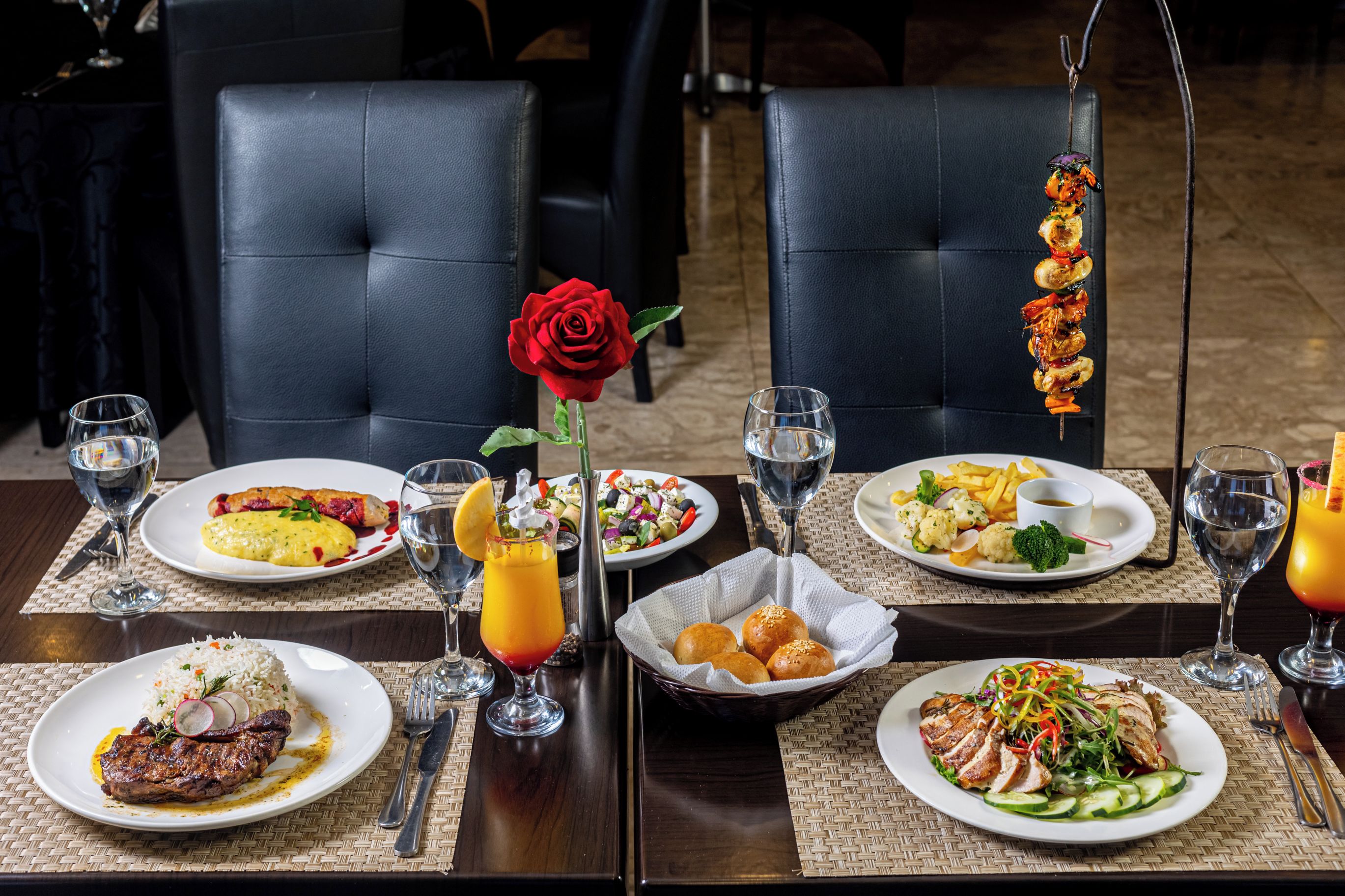 A dining table with various dishes, including steak, salad, and seafood skewers, set on a dark wooden table with woven placemats.