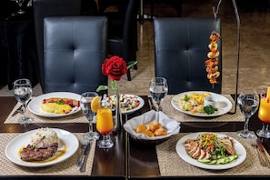 A dining table with various dishes, including steak, salad, and seafood skewers, set on a dark wooden table with woven placemats. - Afrin Prestige (Maputo)
