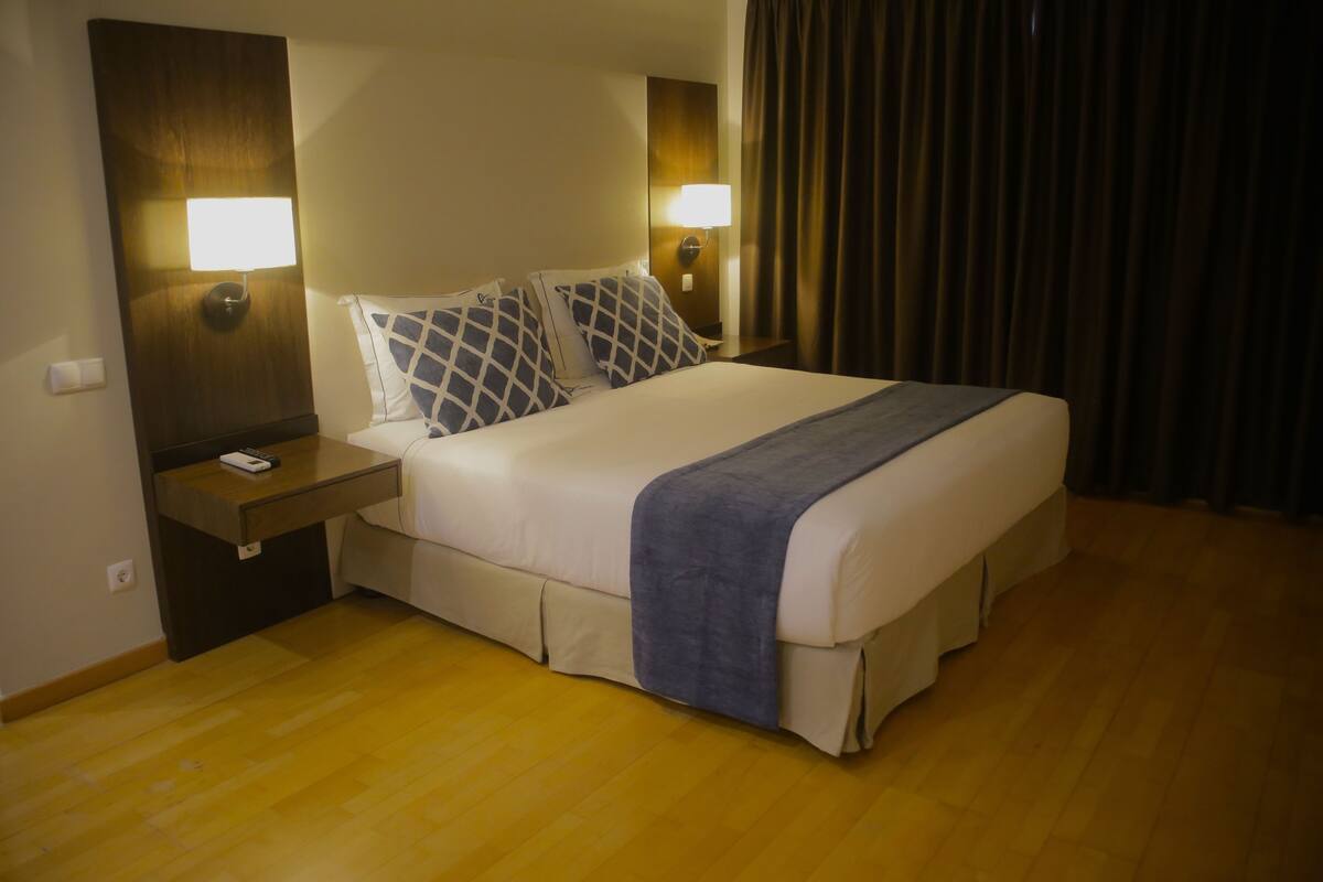 A hotel room with a large bed, two bedside lamps, a nightstand, and a wooden floor.