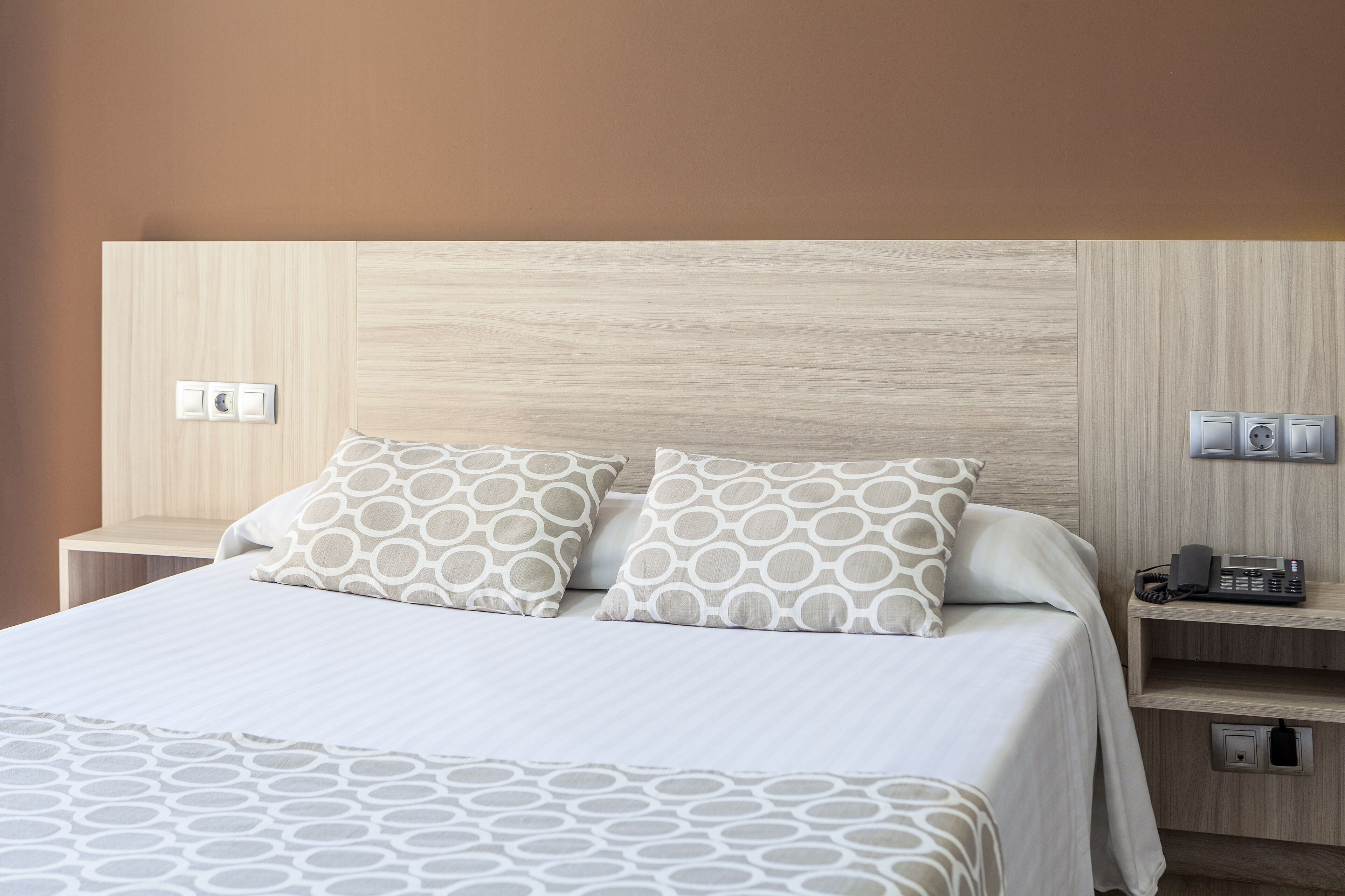 Una cama bien hecha con almohadillas estampadas, un teléfono en la mesita de noche y un interruptor de luz en la pared.