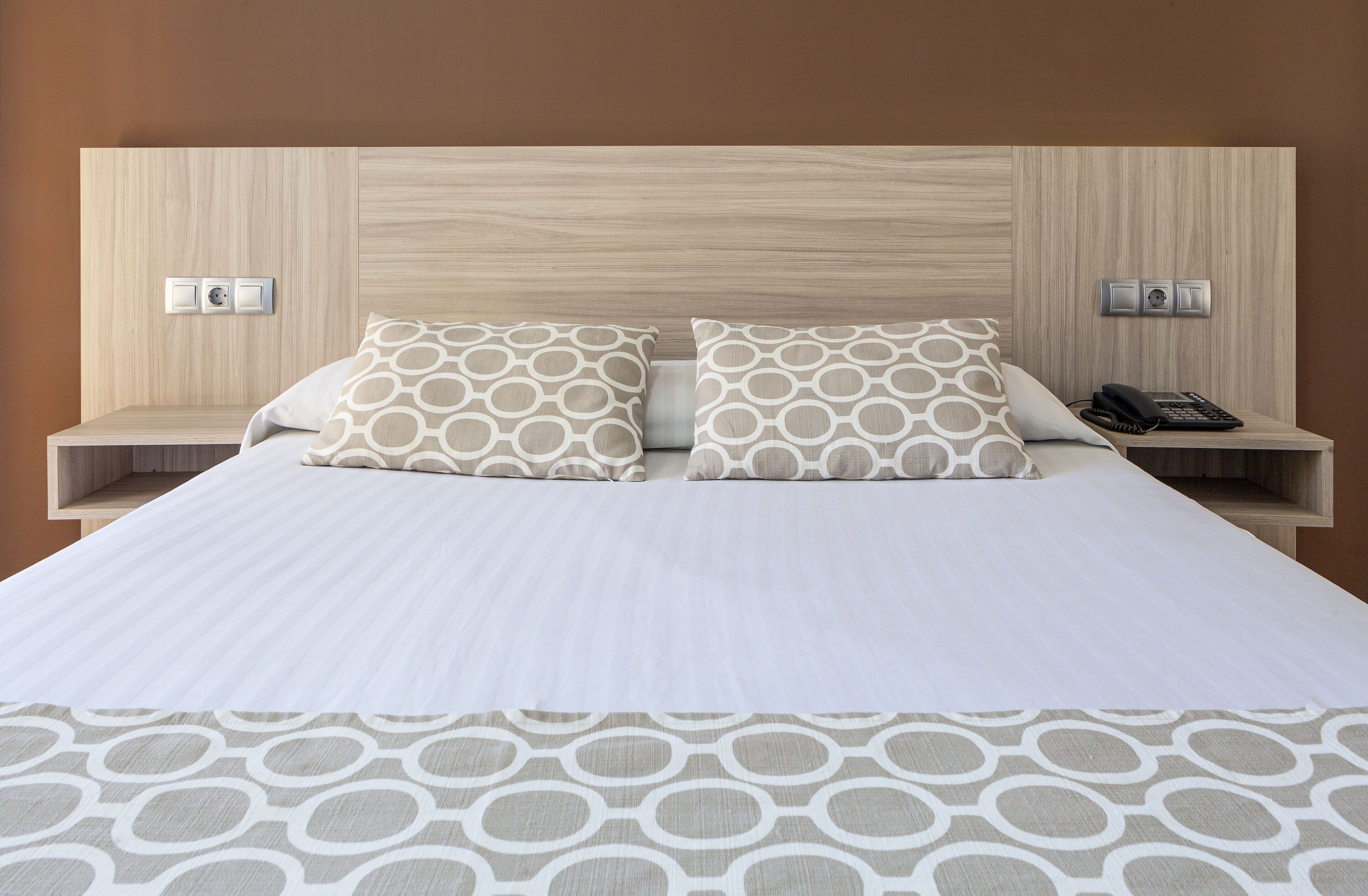 Habitación de hotel con cabecera de madera, dos mesitas de noche, una cama con ropa de cama estampada y un teléfono en una de las mesitas.