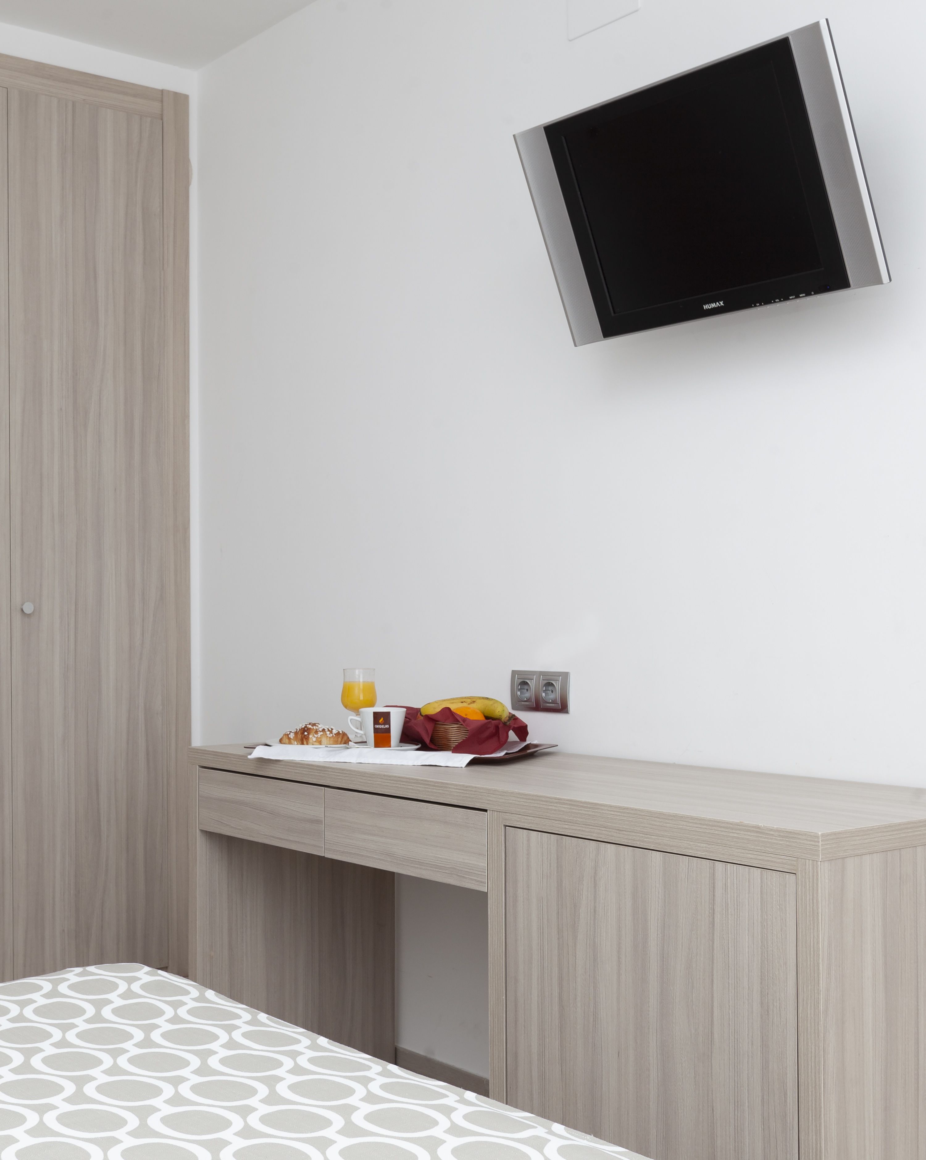 Habitació d'hotel moderna amb un armari de fusta, una taula de treball amb articles per esmorzar i una televisió plana penjada a la paret.