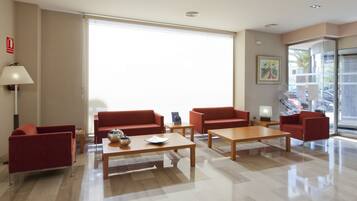 A modern hotel lobby with red sofas, wooden tables, and a large window providing natural light.