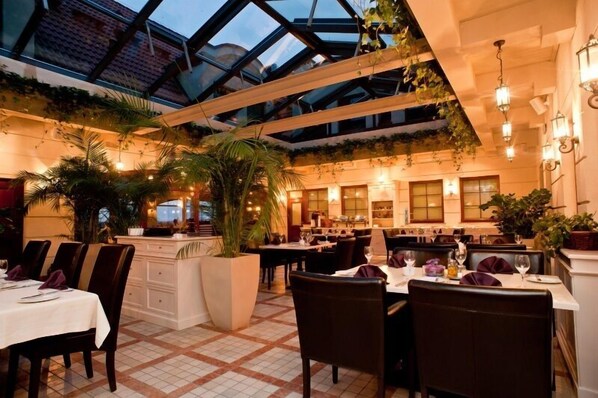 A restaurant with a high ceiling, glass roof, and indoor plants. Tables are set with white tablecloths and dark chairs.