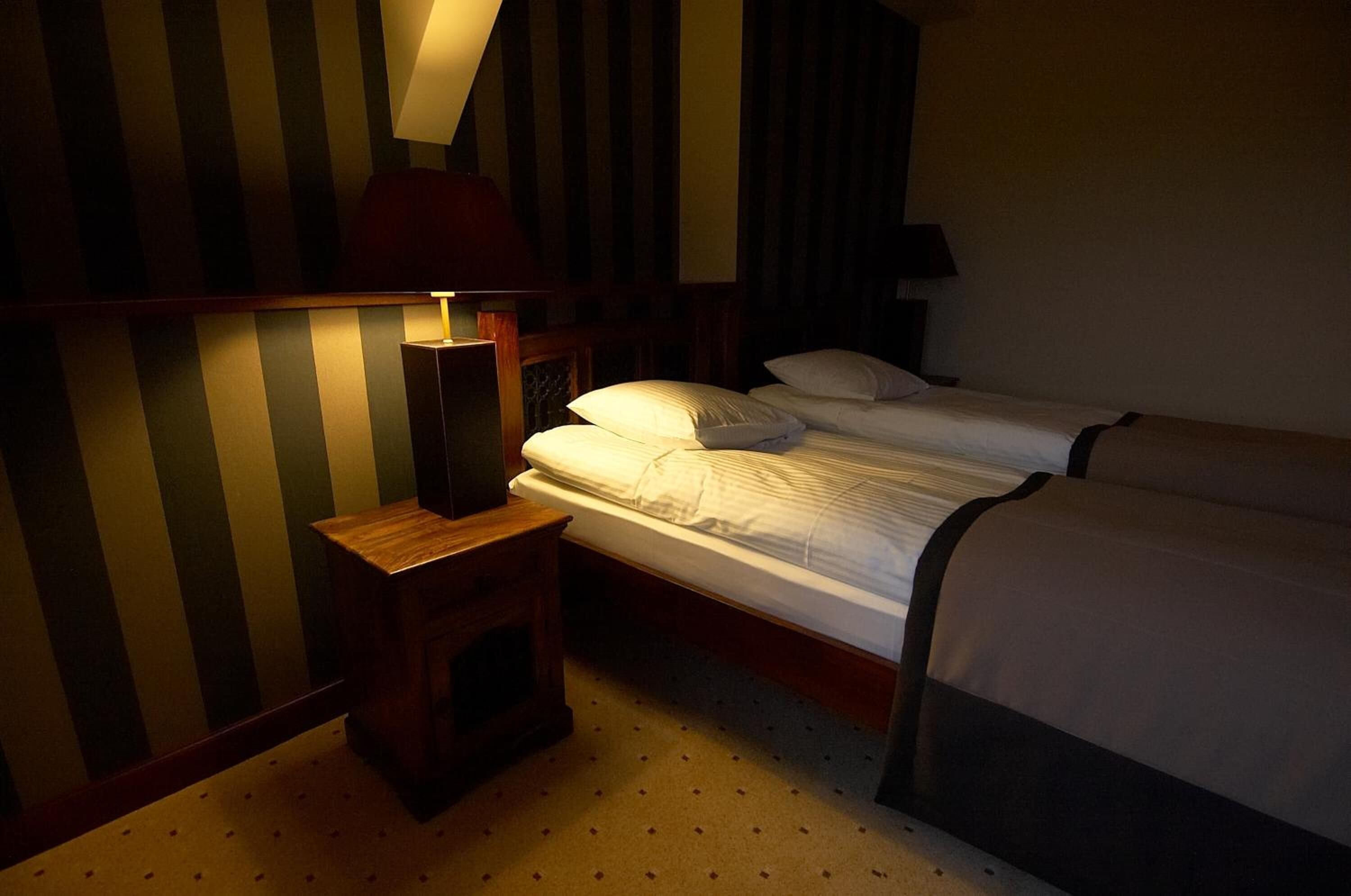 A hotel room with two single beds, a nightstand with a lamp, and striped wallpaper.