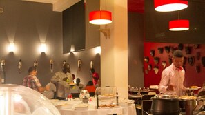 A restaurant with tables set for dining, a chef preparing food, and a wall decorated with various items. - Coimbra Hotel & Spa (Bissau)