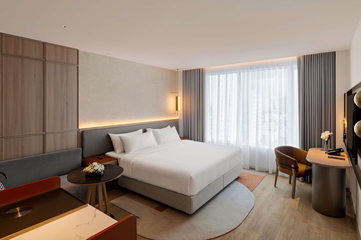 Habitación de hotel moderna con una cama grande, un escritorio, una silla, un televisor y vistas de la ciudad a través de la ventana.