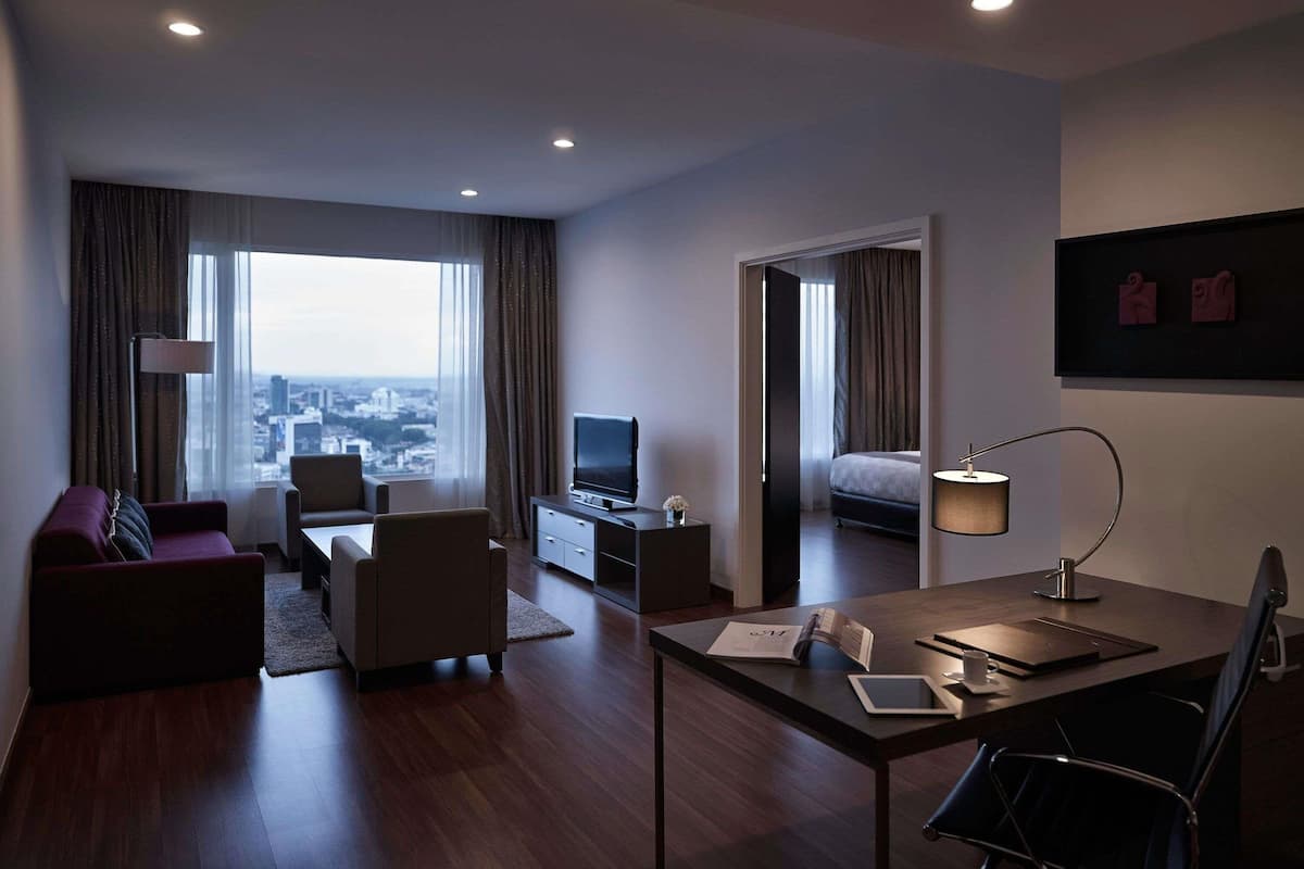 A modern hotel room with a large window, a flat-screen TV, a desk with a lamp, a sofa, and a bed.