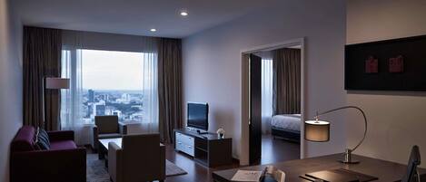 A modern hotel room with a large window, a flat-screen TV, a desk with a lamp, a sofa, and a bed.