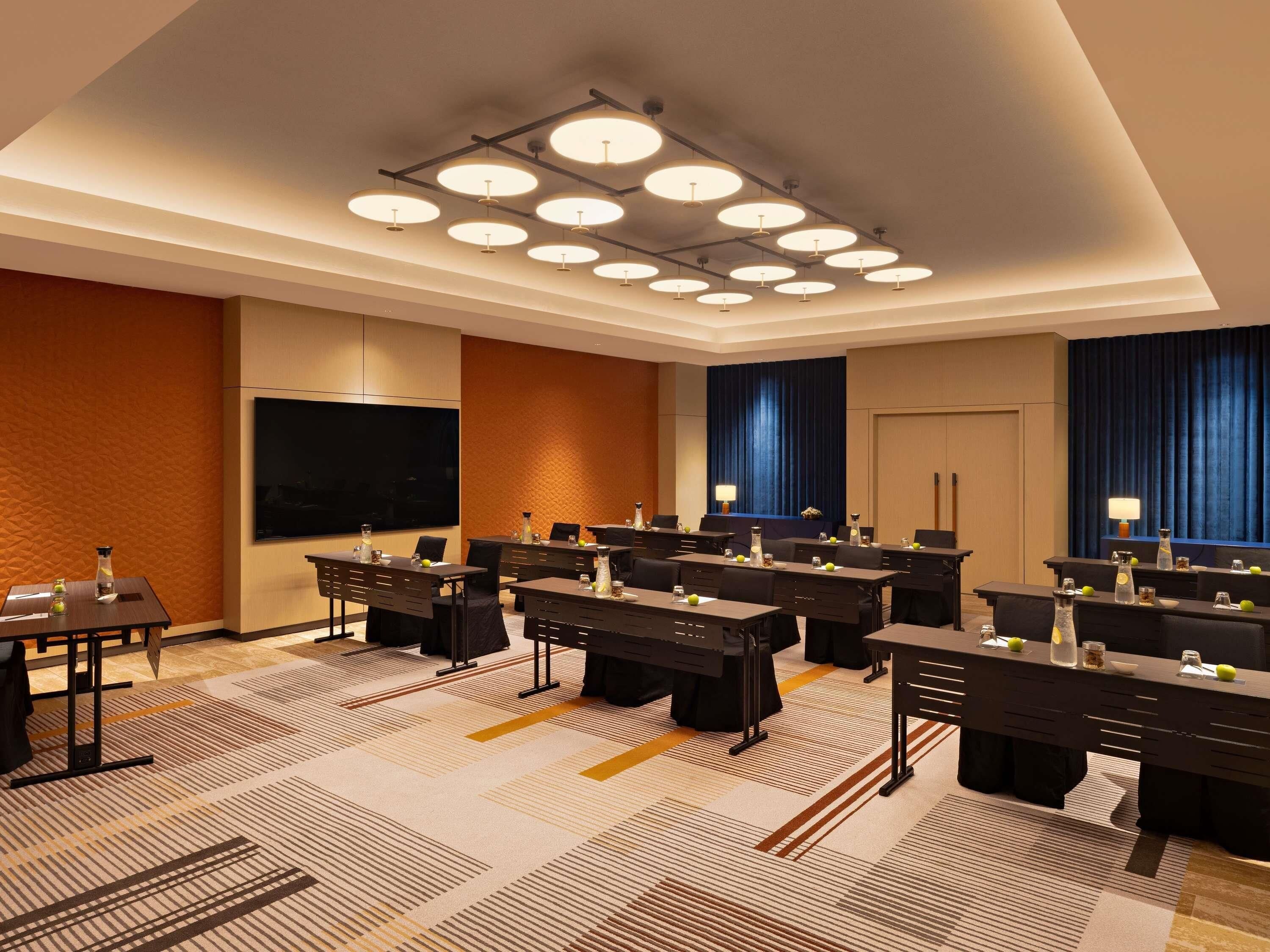A hotel conference room with multiple tables, chairs, a large flat-screen TV, and a ceiling light fixture.