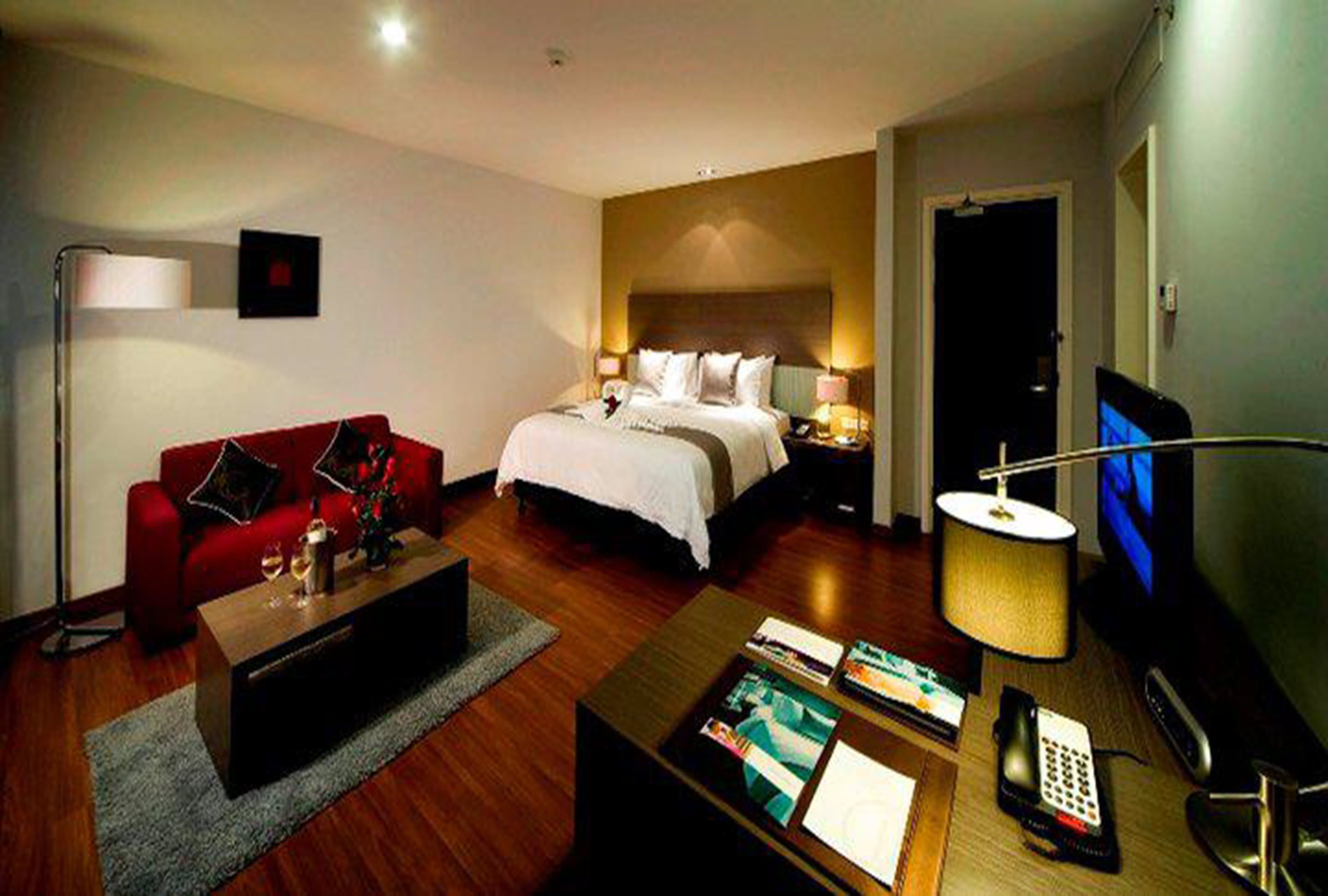 A hotel room with a bed, a red sofa, a coffee table, a TV, and a lamp.