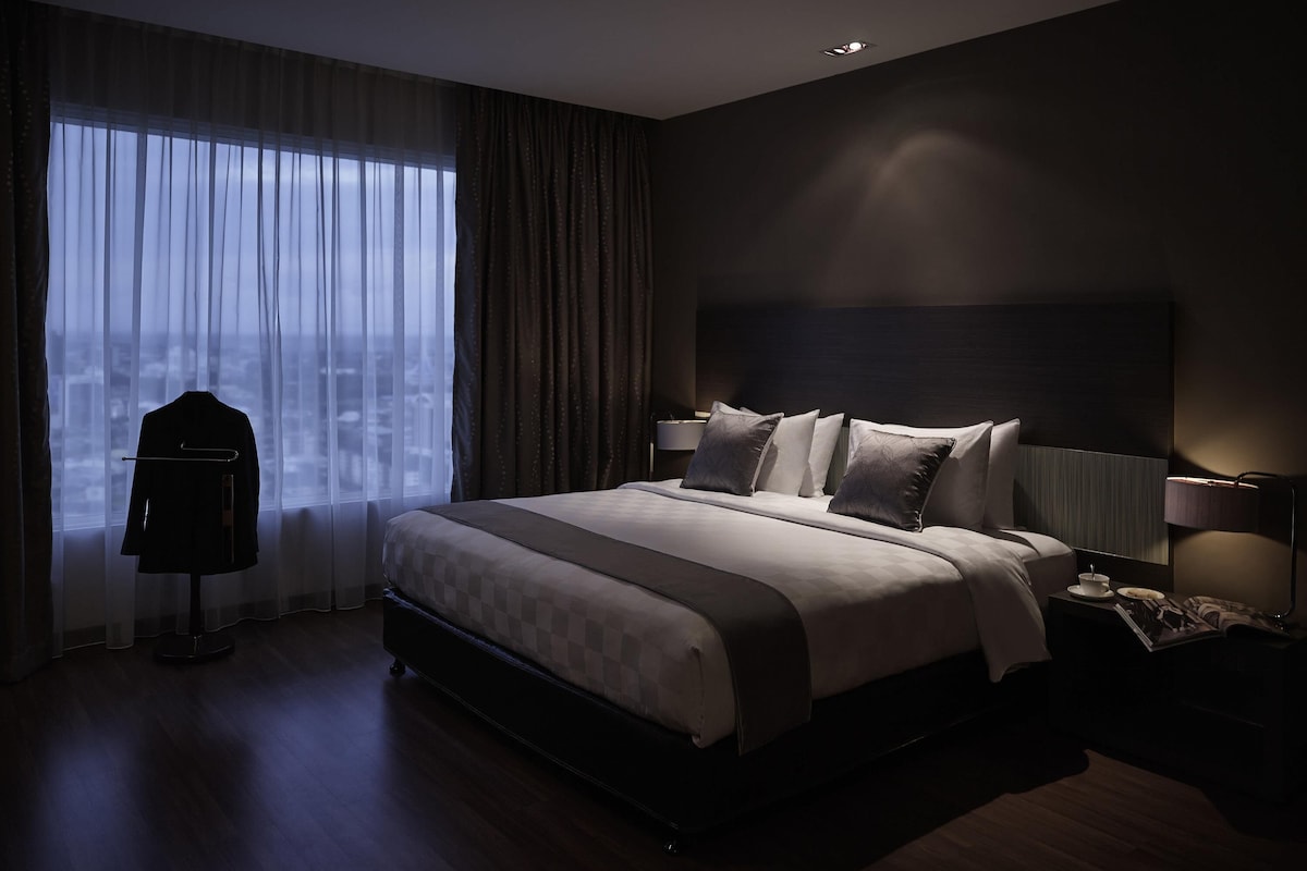 A modern hotel room with a large bed, a nightstand, a lamp, and a view of the city through sheer curtains.
