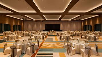 Une vaste salle de banquet, avec de nombreuses tables rondes, chacune recouverte d’une nappe blanche et entourée de chaises, disposées en rangées.
