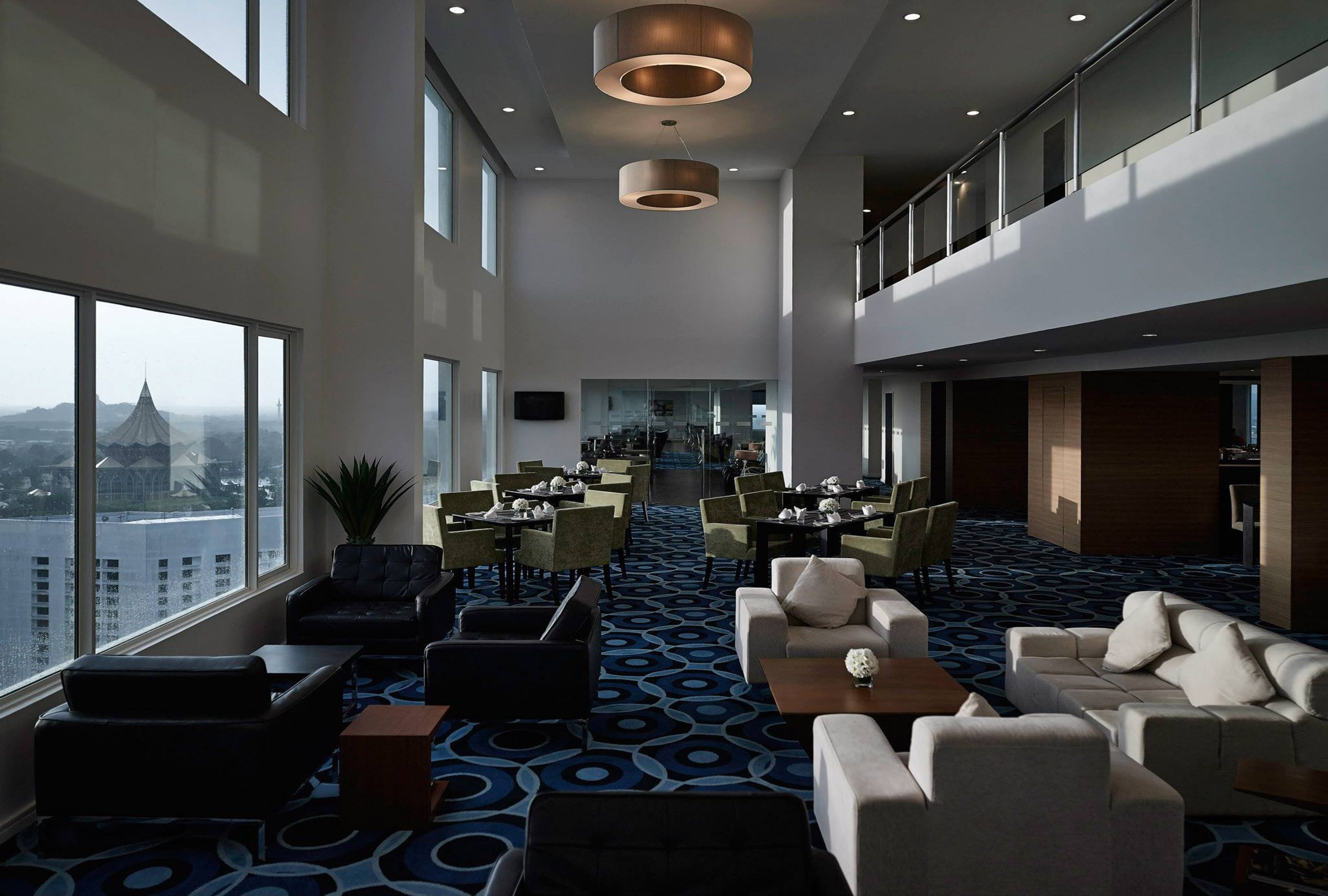 A spacious hotel lobby with high ceilings, large windows offering a city view, and a central seating area with tables and chairs.