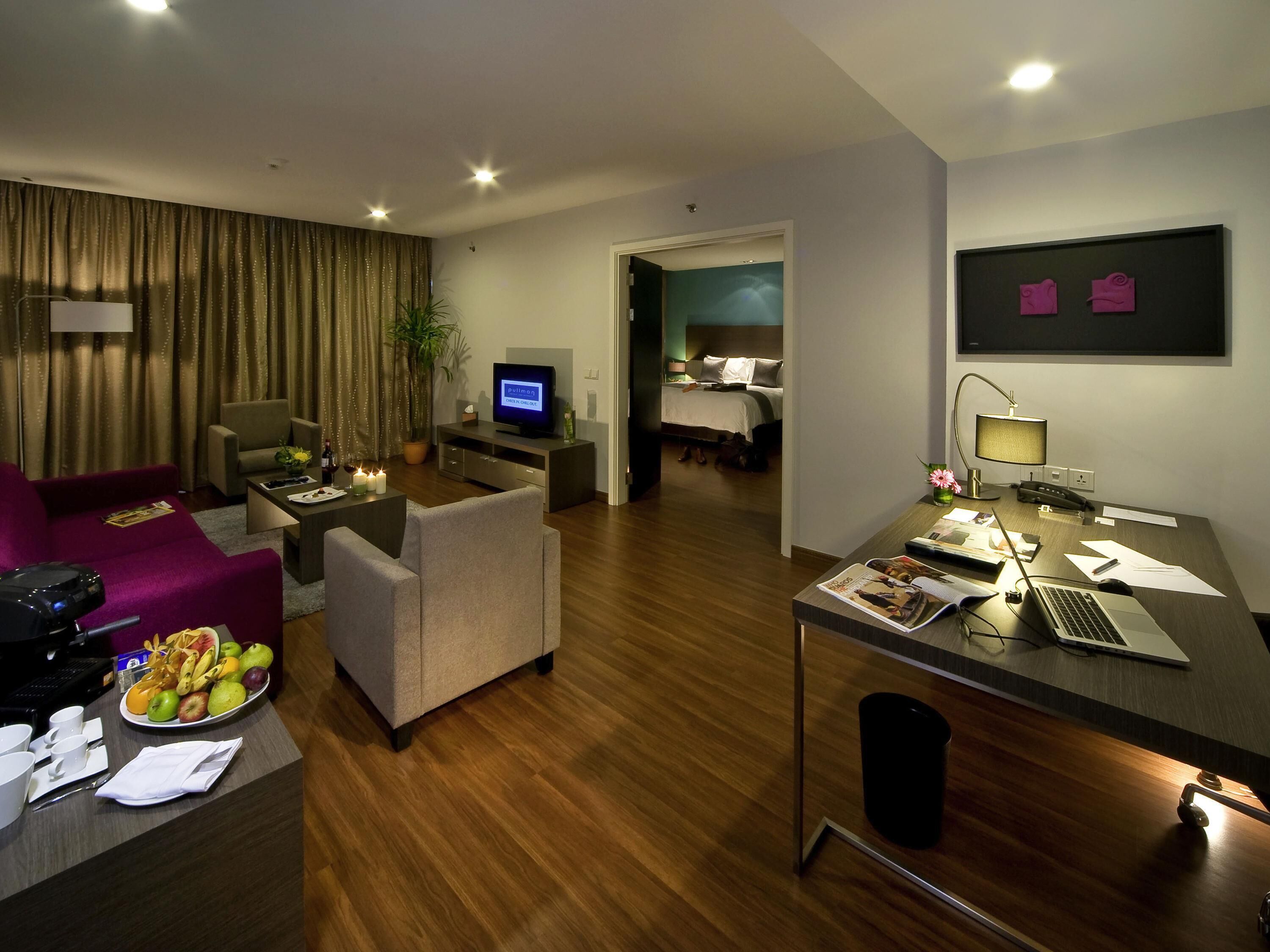 A modern hotel room with a living area, a bedroom, a desk with a laptop, and a television.