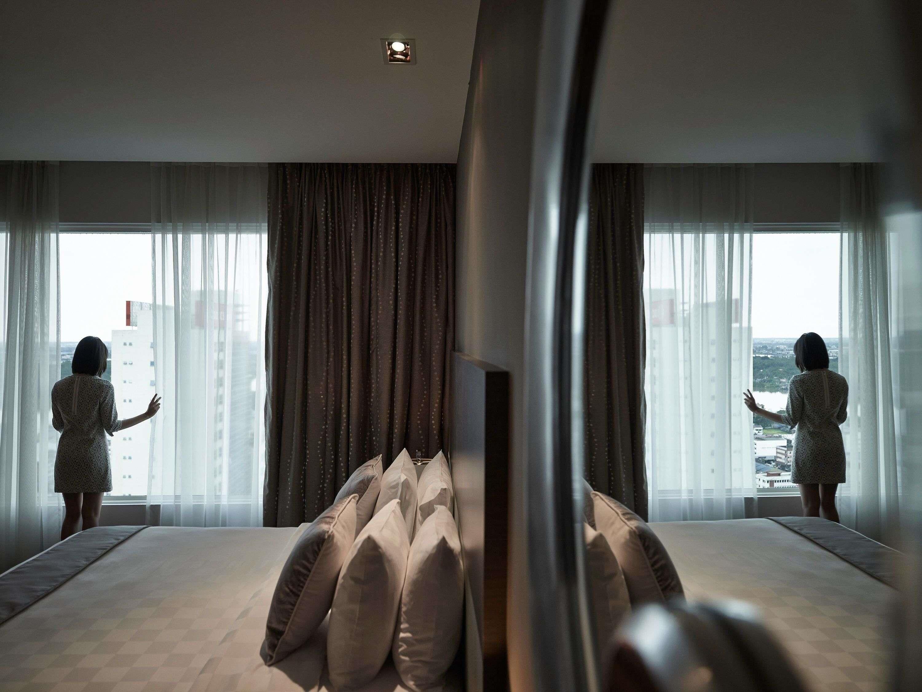 Ein Hotelzimmer mit einem großen Bett, einem Fernseher und Blick auf die Stadt durch transparente Vorhänge.
