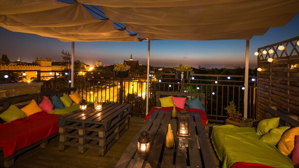 An outdoor seating area with wooden tables and benches, covered by a canopy, and illuminated by string lights. - Hotel Gaja (Poznan)