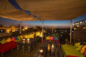 Un área de estar al aire libre con mesas y bancos de madera, cubierta por una marquesina e iluminada con luces colgantes.