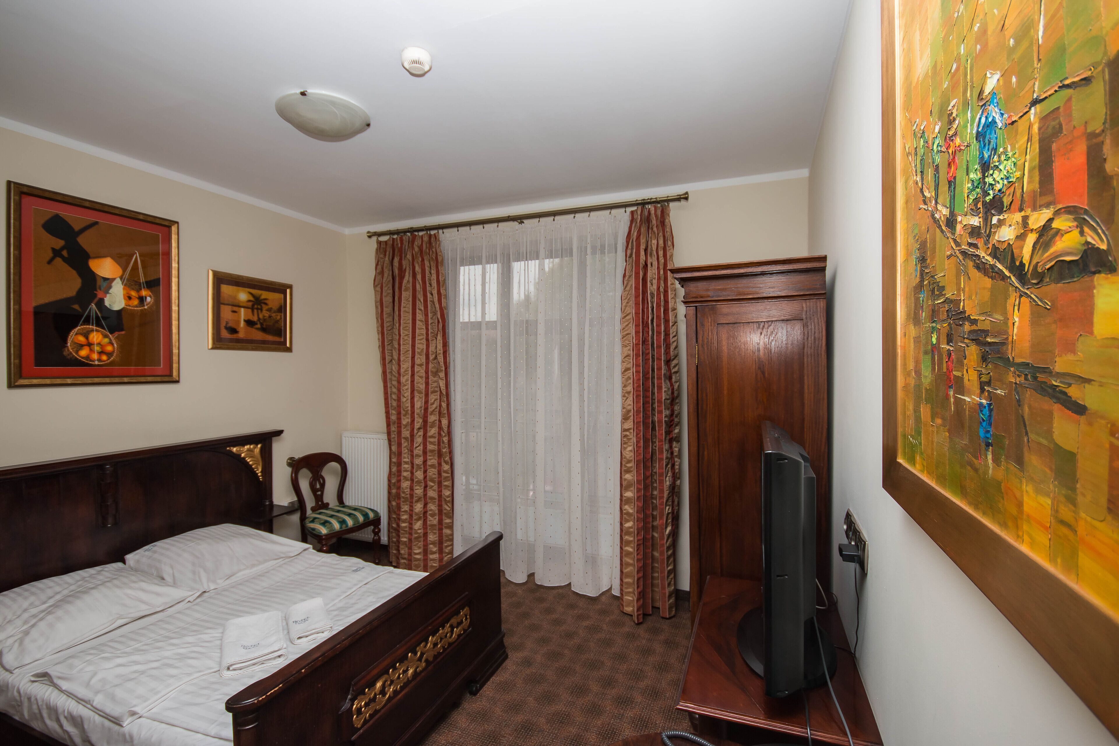 A hotel room with a wooden bed, a desk with a computer, a chair, a window with sheer curtains, and artwork on the walls.