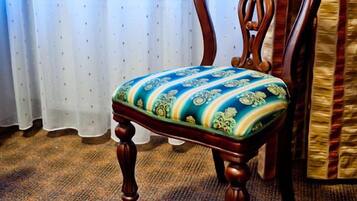 A wooden chair with a patterned cushion in a room with a patterned carpet and a window with sheer curtains.