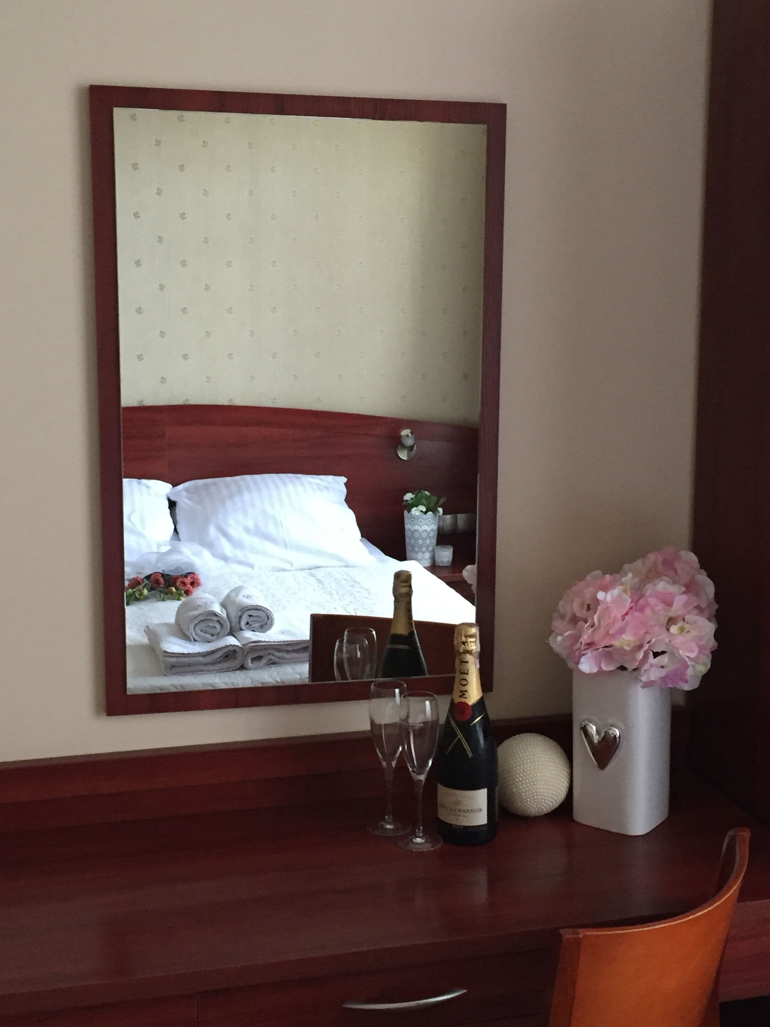 Ein Hotelzimmer mit Bett, Spiegel, einer Champagnerflasche, zwei Gläsern, einer Blumenvase und einem kleinen dekorativen Gegenstand auf einem Holztisch.